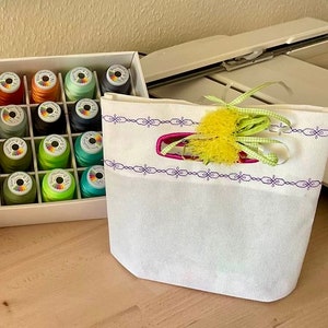 Könnte beinhalten: Eine weiße Stofftasche mit einem lila und weißen Stickmuster. Die Tasche hat ein gelbes und grünes Band, das um die Oberseite gebunden ist. Die Tasche steht auf einem Holztisch neben einer Schachtel mit bunten Garnrollen.