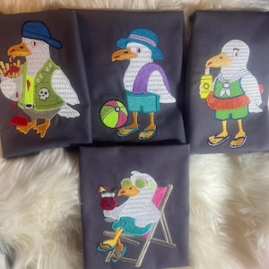 Könnte beinhalten: Bestickte graue Stoffquadrate mit Cartoon-Möwen in Strandoutfits. Eine Möwe trägt einen Hut und eine Weste, eine andere hält einen Strandball und eine dritte hat eine Schwimmweste. Die letzte Möwe entspannt sich in einem Strandstuhl.