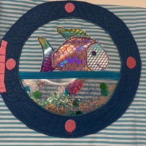 Könnte beinhalten: Ein gestickter Fisch in Blau und Rosa in einem blauen Bullauge mit Glitzer im Inneren.