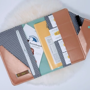 Könnte beinhalten: Ein roségoldener Reißverschlussordner mit einer grauen und weißen Sternenmuster-Stoffauskleidung. Der Ordner hat mehrere Taschen und Fächer zum Aufbewahren von Dokumenten und Karten. Der Text "MUTTERPASS" und "Feststellung der Hörfähigkeit" ist im Inneren des Ordners sichtbar.