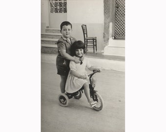 Children play with the bicycle. Nikaia, Greece 1960s. Vintage photo postcard size. Photographer: Manolis. [53006]