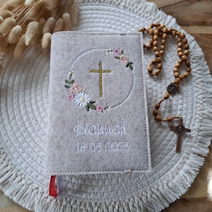 Könnte beinhalten: Ein grauer Filzbuchumschlag mit einem weißen gestickten Kreuz und einem Blumendesign. Der Umschlag ist mit dem Namen "Hannah" und dem Datum "18.05.2025" personalisiert.