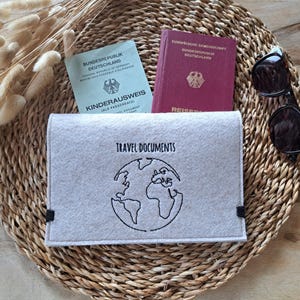 Könnte beinhalten: Ein grauer Filz-Reisepass-Etui mit schwarzen Nähten und dem aufgenähten Text "TRAVEL DOCUMENTS" auf der Vorderseite. Unter dem Text ist ein schwarz-weißes Globus-Motiv gestickt.