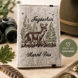 Könnte beinhalten: Ein grauer Filz-Passhalter mit gesticktem Hirsch und Waldszene. Der Text "Jagdschein" und ein Name sind auf die Vorderseite gestickt. Oben auf dem Halter steht "PERSONALISIERBAR" und "MIT DEINEM NAMEN".