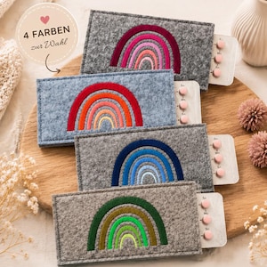 Könnte beinhalten: Vier rechteckige Pillenhüllen aus grauem Filz mit gestickten Regenbogen-Motiven in Pink, Orange, Blau und Grün. Jede Hülle enthält einen Blister mit Tabletten. Der Text lautet PILLENHÜLLE MIT REGENBOGEN-STICKEREI, SÜSS, PRAKTISCH, HANDGEMACHT, 4 FARBEN zur Wahl, LIEBEVOLLE STICKEREI, SCHÜTZT DEINE PILLE DISKRET, WEICHES FILZMATERIAL, PERFEKT ALS GESCHENK, Dein täglicher Begleiter mit Stil & Herz, UnVergleichlichesDE.