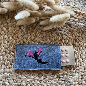 Könnte beinhalten: Ein grauer Filzbeutel mit einem rosa gestickten Feenmotiv. Der Beutel liegt neben einer Blisterpackung mit rosa Pillen.