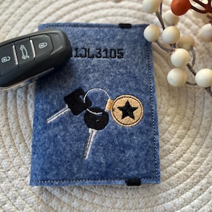 Könnte beinhalten: Ein blauer Schlüsselanhänger aus Filz mit einem schwarzen gestickten Design aus zwei Schlüsseln und einem Stern. Der Text "1JL3105" ist auf der Vorderseite des Schlüsselanhängers gestickt.