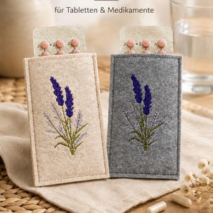 Könnte beinhalten: Zwei rechteckige Pillenhüllen aus Filz, eine beige und eine graue, jeweils mit einer gestickten Lavendelblüte. Jede Hülle enthält einen Blister mit rosa Tabletten. Der Text lautet Pillenhüllen, PRAKTISCH STILVOLL HANDLICH, für Tabletten & Medikamente, 5,5 x 10 cm.