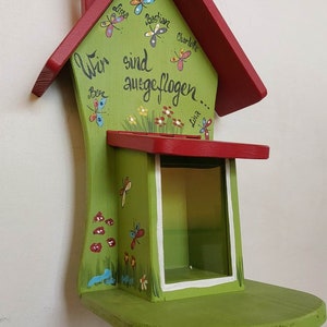 Könnte beinhalten: Ein grünes Holzvogelhaus mit rotem Dach und einem kleinen Fenster. Das Vogelhaus ist mit Schmetterlingen und Blumen verziert. Der Text "Wir sind ausgeflogen..." ist auf die Vorderseite des Vogelhauses gemalt.