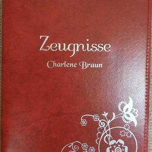 Puede incluir: Un archivador de cuero rojo con el título "Zeugnisse Charlene Braun" grabado en letras plateadas. El archivador tiene un diseño floral plateado en la esquina inferior derecha.