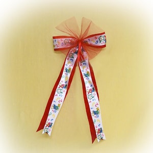 May include: A red bow with white ribbon featuring colourful numbers and cartoon characters. The ribbon is tied with a sheer red mesh.