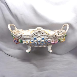 以下が含まれることがあります： 金色のトリムと花のアクセントが付いた白い磁器製のセンターピースボウル。ボウルは、青、ピンク、黄色の花が咲く格子模様のデザインです。2つのハンドルがあり、3つの小さな足の上に置かれています。