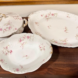 Może przedstawiać: Zestaw antycznych białych porcelanowych naczyń do serwowania z kwiatowym wzorem w kolorze różowym i zielonym. Zestaw zawiera pokrytą wazę do zupy, duży owalny półmisek i okrągłą miskę do serwowania z falistymi krawędziami i złotym wykończeniem.