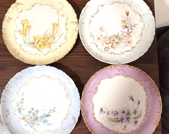 Pode incluir: Quatro pratos redondos de cerâmica com bordas recortadas e acabamento dourado. Cada prato apresenta uma cor de borda pastel diferente: amarelo, azul claro, azul pálido e lavanda. Cada prato exibe uma ilustração floral única no centro.