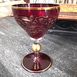 May include: A deep red glass goblet featuring a gold rim and intricate gold floral etchings on the bowl. The stem includes a central gold sphere accent, and the circular base is finished with a thin gold border, creating a cohesive design.