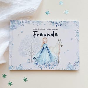 Op de afbeelding: Een rechthoekig boek met een witte kaft voorzien van een blauwe winterse illustratie van een persoon in een japon naast een wit paard. De tekst "Meine liebsten & zauberhaftesten Freunde" staat in het zwart op de voorzijde gedrukt.
