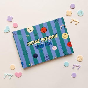 Könnte beinhalten: Ein rechteckiges Buch mit einem blau-grün gestreiften Einband, verziert mit verschiedenen bunten Smiley-Symbolen und dem Text "MEINE FREUNDE UND ICH" in Gelb und Blau. Um das Buch herum sind kleine pastellfarbene Dekorationselemente verteilt.