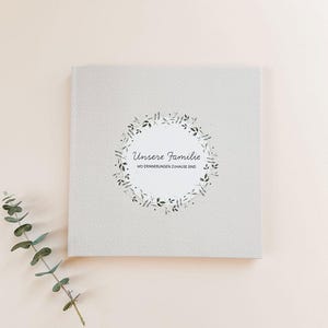 Könnte beinhalten: Ein graues Journal mit einem weißen Kreis und einem schwarzen floralen Design. Der Text "Unsere Familie" und "Wo Erinnerungen Zuhause Sind" ist auf dem Cover gedruckt.