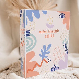 Könnte beinhalten: Ein Ordner mit einem farbenfrohen Design mit dem Text "MEINE SCHULZEIT AMIRA". Das Cover hat einen hellrosa Hintergrund mit blauen, orangefarbenen und lila abstrakten Formen sowie einem Gittermuster und gepunkteten Akzenten.