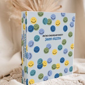 Könnte beinhalten: Eine hellblaue Box mit gelben, grünen, blauen und lila Smileys. Der Text "MEINE KINDERGARTENZEIT JAKOB GIESSEN" ist in Schwarz gehalten. Die Box liegt auf einem weißen Tuch.