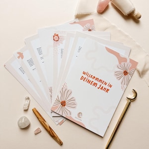 Könnte beinhalten: Eine Sammlung weißer Karten mit floralen Designs und dem Text "Willkommen in Deinem Jahr" in Orange. Die Karten sind mit einem goldenen Stift, Kristallen und einem Holzstab auf einer cremefarbenen Oberfläche angeordnet.