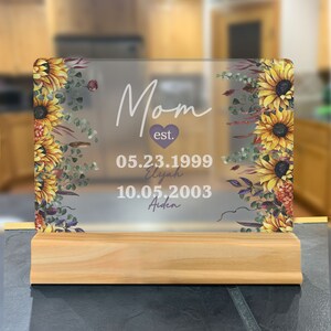 May include: A clear acrylic sign with a wood stand featuring a floral design with sunflowers and greenery. The sign reads "Mom est. 05.23.1999 Elijah 10.05.2003 Aiden".