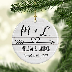May include: White ceramic Christmas ornament with a woodgrain design. The ornament features the text "M + L" with an arrow and heart design. The text "Melissa & Landon" and "December 10, 2019" is also included.