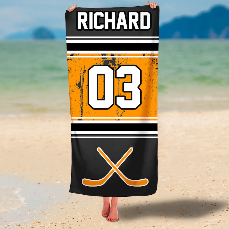 May include: Black beach towel with orange and white hockey jersey design. The towel features the name "RICHARD" and the number "03" in white on an orange background. Two crossed hockey sticks are at the bottom of the towel.