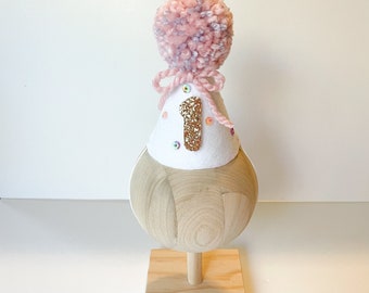 Puede incluir: Un gorro de fiesta de cumpleaños hecho a mano, sobre una base de madera. El gorro blanco presenta el número "1" en purpurina dorada y lentejuelas de colores. Está rematado con un pompón rosa y morado y atado con un lazo rosa.
