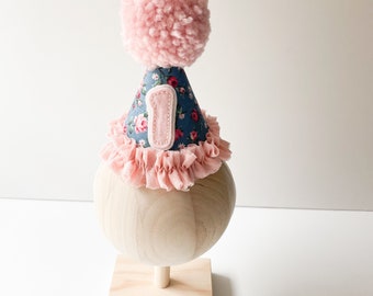 May include: A festive party hat with a blue floral pattern, a pink pom-pom, and a pink ruffle trim. The hat features a felt number "1" applique, perfect for a first birthday celebration. The hat is displayed on a wooden stand.