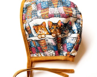 May include: A baby bonnet with a patchwork pattern in shades of blue, pink, and yellow. The bonnet features a print of three kittens sleeping on a pillow. The trim and ties are a mustard yellow color.