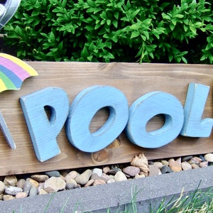 Könnte beinhalten: Ein hölzernes Pfeilschild mit dem Wort "POOL" in blauen Buchstaben, einem bunten Strandschirm und grünen Flip-Flops. Das Schild steht auf einem Bett aus kleinen Steinen.