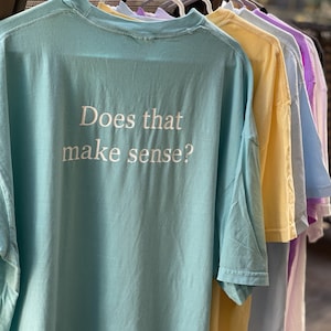 May include: A light blue t-shirt with the text "Does that make sense?" printed in white on the back. The shirt is hanging on a rack with other t-shirts in various colors.
