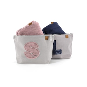 Könnte beinhalten: Zwei Canvas-Beutel, einer mit einem großen rosa "S" und dem Namen "Sina", der andere mit einem blauen "L" und dem Namen "Lenny". Jeder Beutel enthält ein gefaltetes Tuch. Die Beutel sind hellgrau mit braunen Lederetiketten.