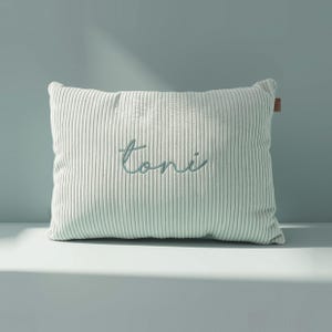 May include: A rectangular light green corduroy pillow featuring a vertical ribbed texture. The name "toni" is embroidered in a dark teal cursive script across the center of the pillow. A small brown rectangular tag is visible on the right side.