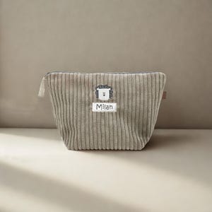 Pode incluir: Uma bolsa com fecho de correr em veludo cotelê cor de toupeira com textura de canelado vertical. No centro da frente, um ícone de cara de leão branco bordado sobre um remendo de tecido branco com o nome Milan em letras pretas.