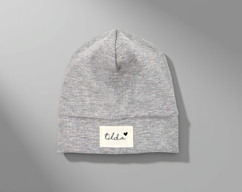 May include: A light grey ribbed knit beanie featuring a rectangular off-white fabric patch on the front. The patch displays the word tilda in a black cursive script font, accompanied by a small black heart symbol positioned to the right.