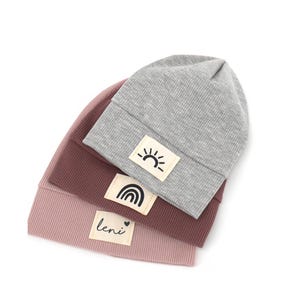 Könnte beinhalten: Drei gerippte Strickmützen in Grau, Mauve und Staubrosa. Jede Mütze hat einen rechteckigen Aufnäher mit einer Grafik: eine Sonne, ein Regenbogen und der Name "leni".