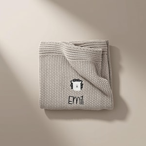 May include: A beige knitted blanket featuring a textured garter stitch pattern. The blanket is embroidered with a white and black lion face graphic and the name EMIL in black thread, positioned centrally on the folded fabric surface.