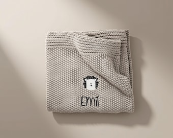 Op de afbeelding: Een beige gebreide deken met een gestructureerd ribbelsteekpatroon. De deken is geborduurd met een wit en zwart leeuwengezicht en de naam EMIL in zwarte draad, centraal geplaatst op het oppervlak van de gevouwen stof.
