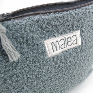 May include: A close-up of a textured, blue-grey pouch with a zipper closure. The pouch has a small, rectangular label with the name "Malea" embroidered on it. A grey tassel hangs from the zipper pull.