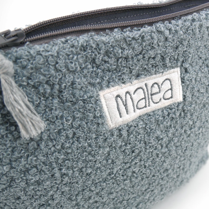 May include: A close-up of a textured, blue-grey pouch with a zipper closure. The pouch has a small, rectangular label with the name "Malea" embroidered on it. A grey tassel hangs from the zipper pull.