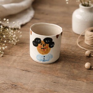 May include: A white ceramic mug with a hand-painted face design. The face has black hair, a tan face, and a blue collar. Brown accents are around the rim. The mug is on a wooden surface.