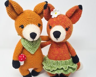 May include: Two crocheted fox plushies, one wearing a green skirt and the other a green bandana. Both have black eyes and noses. There are also crocheted mushrooms and flowers in the image.