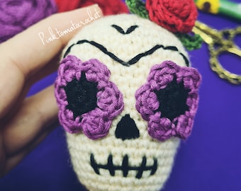May include: A crocheted sugar skull with purple flowers for eyes, a black stitched mouth, and a red flower on top. The skull is cream colored and has black stitching for the nose and eyebrows. The skull is held in a hand against a purple background.