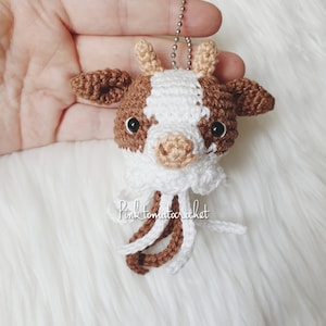 May include: A handmade crochet cow keychain with brown and white details, including a small bell chain. The cow's head features black eyes and a white muzzle. The keychain has a white and brown fringe. The text "Pink tomatocrochet" is visible.