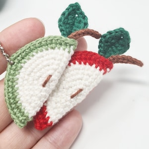 Pode incluir: Pingente de fatia de maçã de crochê, com fio branco, vermelho e verde. A maçã tem casca verde, polpa branca e miolo vermelho. Fio marrom para o caule e folhas. O pingente é preso a uma corrente prateada.