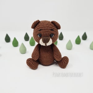 May include: A brown crocheted teddy bear with black eyes and a white nose sitting on a white surface. The bear is surrounded by small green wooden shapes.