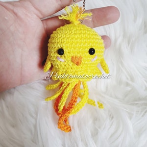 May include: A yellow crocheted chick keychain with orange yarn for the legs and a small silver chain attached to the top. The chick has black eyes and a small orange beak.