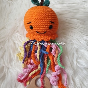 May include: A crocheted orange octopus toy with a wooden ring. The octopus has a smiling face and rainbow colored tentacles.